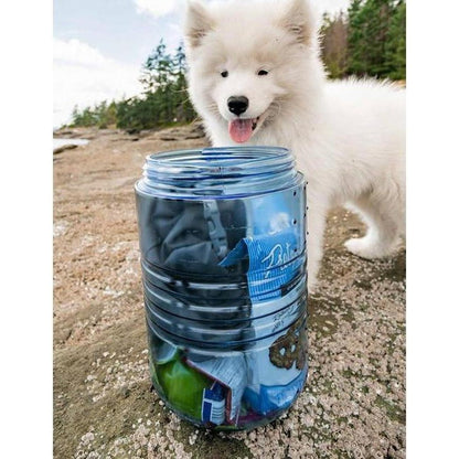 A fluffy white Samoyed puppy stands behind a blue Bear Vault BV500 - Journey food storage container on a rocky outdoor trail.