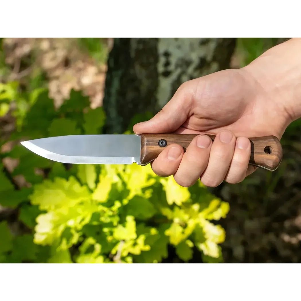 A hand holds the Beavercraft Breeze Bushcraft knife with a wooden handle, set against green foliage and a tree trunk.
