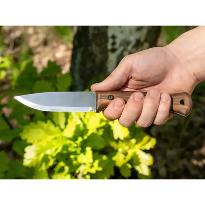 A hand holds the Beavercraft Breeze Bushcraft knife with a wooden handle, set against green foliage and a tree trunk.
