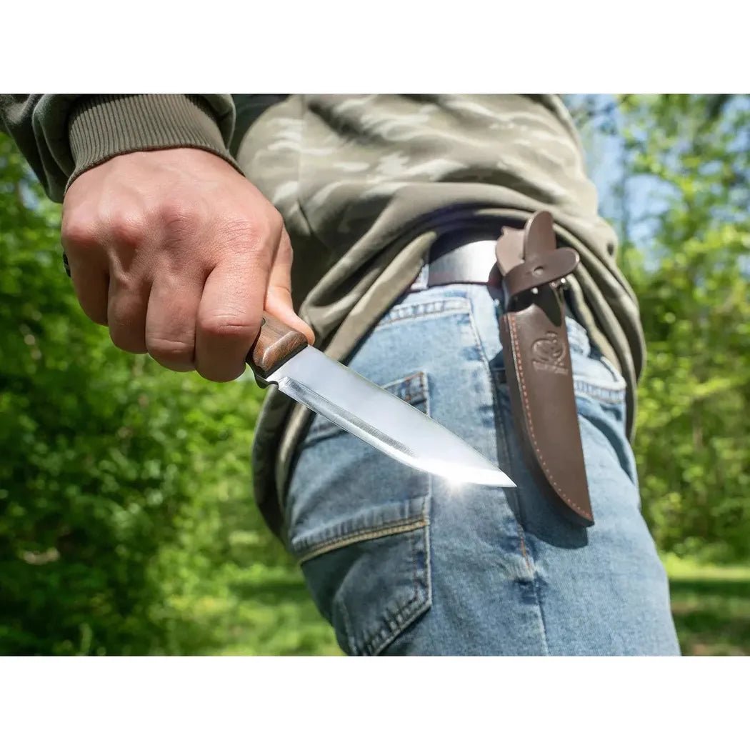 A person holds the Beavercraft Breeze Bushcraft knife with its brown sheath attached to their waistband, set against a green outdoor background.