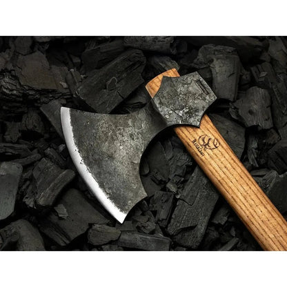 A side view of the Beavercraft Chopping Hewing Axe with a wooden handle, displayed on a bed of charcoal-colored wood chips.