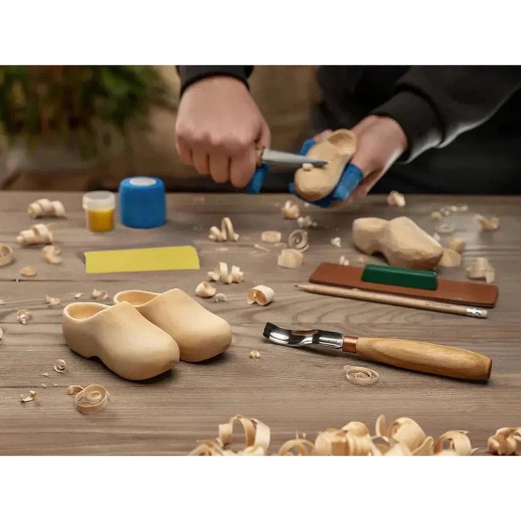 A person carves a wooden clog with the Beavercraft Clogs Carving Kit on a workbench with finished clogs and wood shavings.