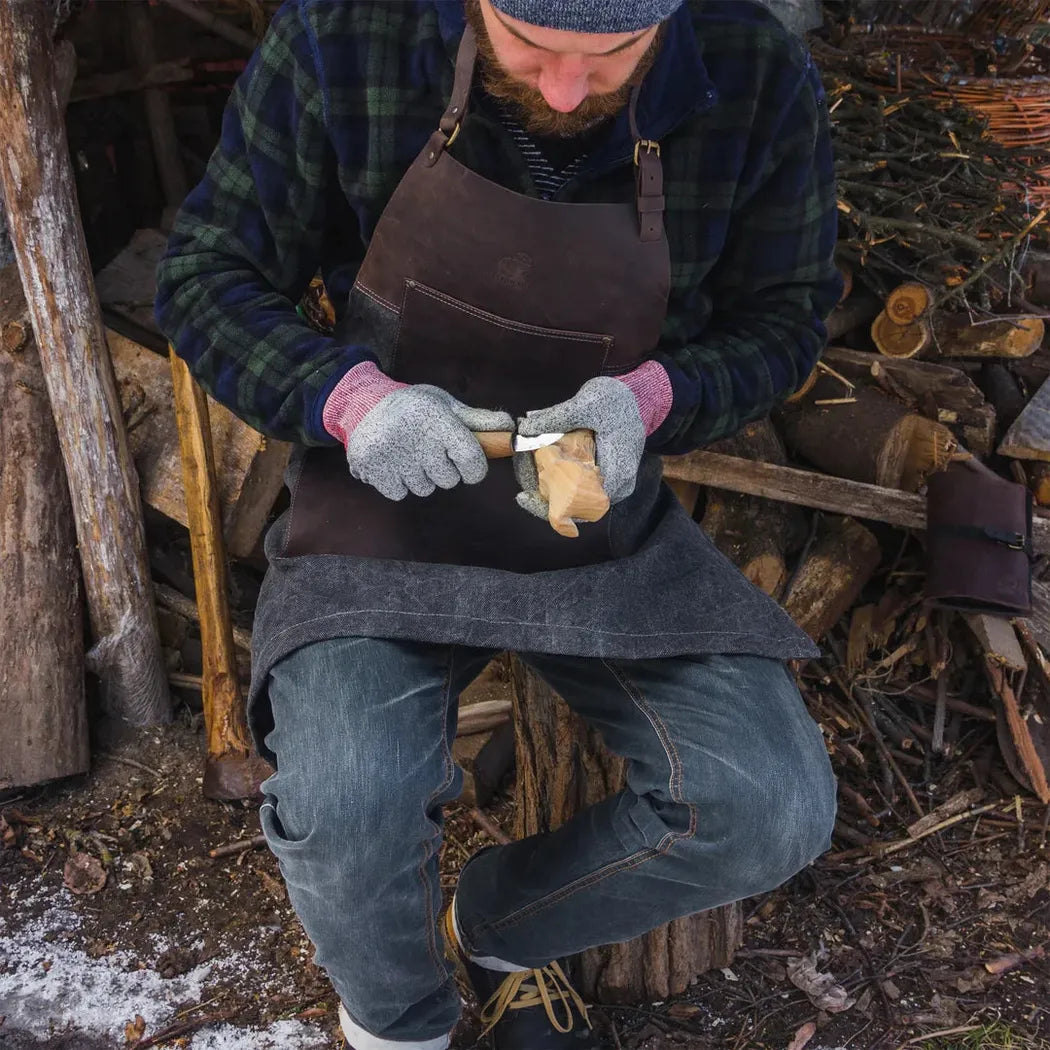 A man in a plaid shirt and jeans wears the Beavercraft Cowhide Apron Leather while carving wood outdoors.