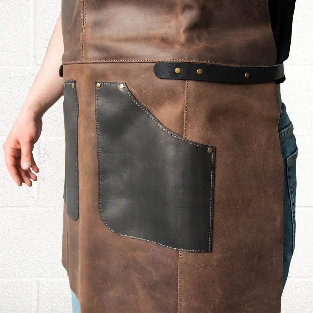 A front view of the Beavercraft Cowhide Apron Leather with a charcoal pocket and black belt, worn by a person in blue jeans against a white brick wall.