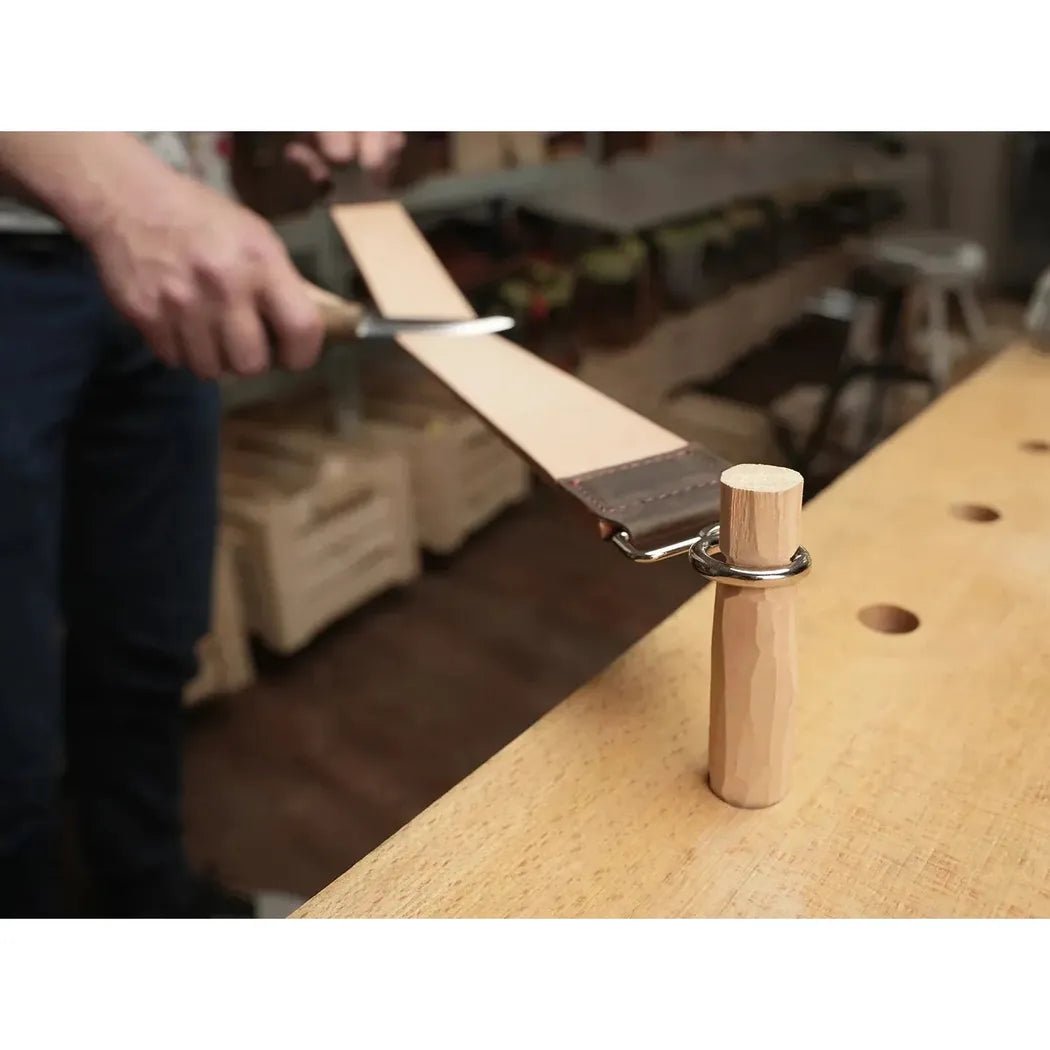 A person uses the Beavercraft Double-Sided Leather Hanging Strop on a workbench with a wooden post and metal ring.