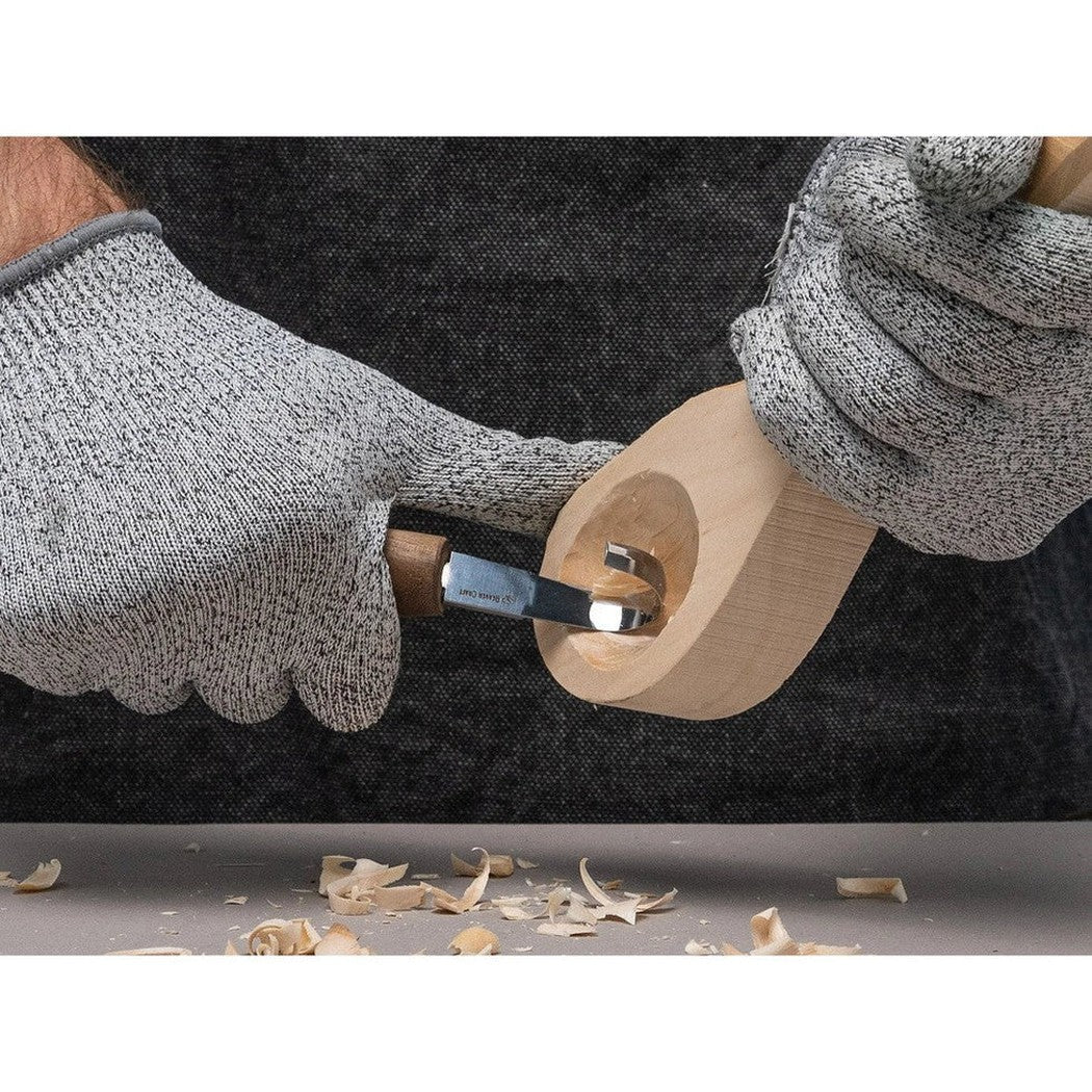 A person wearing gray gloves uses the Beavercraft Luxury Spoon Carving Set With Walnut Handles to carve a wooden spoon, with wood shavings on the table below.