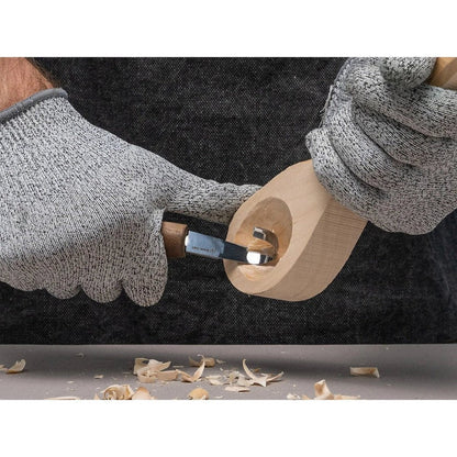 A person wearing gray gloves uses the Beavercraft Luxury Spoon Carving Set With Walnut Handles to carve a wooden spoon, with wood shavings on the table below.