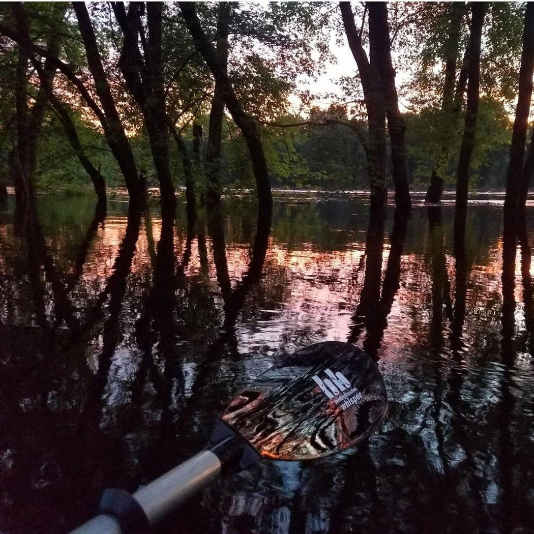 A close-up of the Bending Branches Whisper AS paddle in Black, showing the blade and handle, with a serene river and tree reflections in the background.