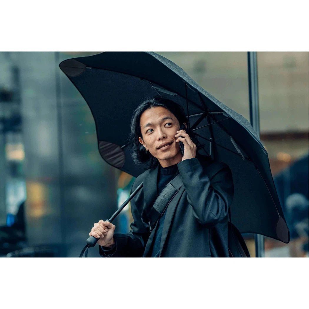 A man in a black coat holds a Black Blunt Classic umbrella while talking on his phone in an urban setting.