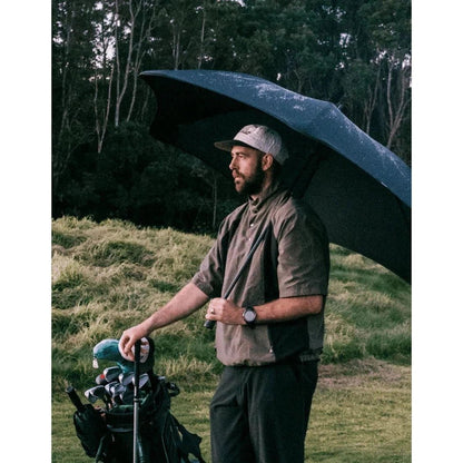 A man stands on a grassy golf course holding a Black Blunt Sport umbrella over his head.