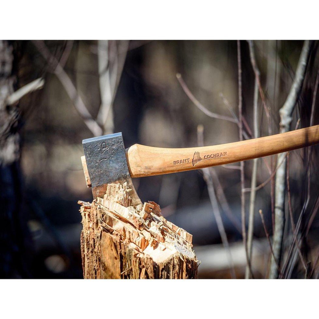 A close-up of the Brant & Cochran Allagash Cruiser Maine Wedge Camp Axe with its wooden handle and metal head resting on a tree stump in a wooded background.