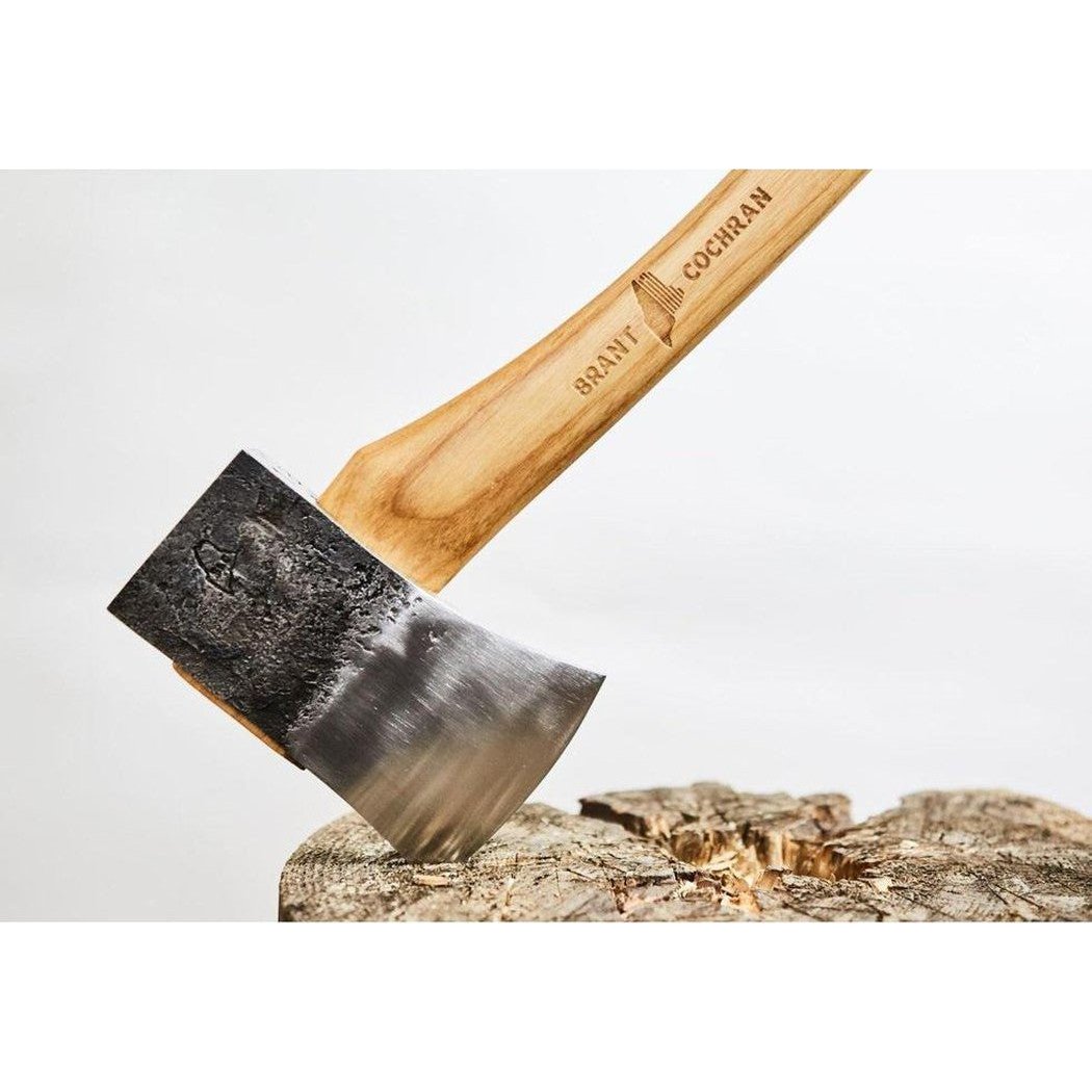 A close-up of the Brant & Cochran Allagash Cruiser Maine Wedge Camp Axe with its wooden handle and charcoal-colored metal head resting on a tree stump.