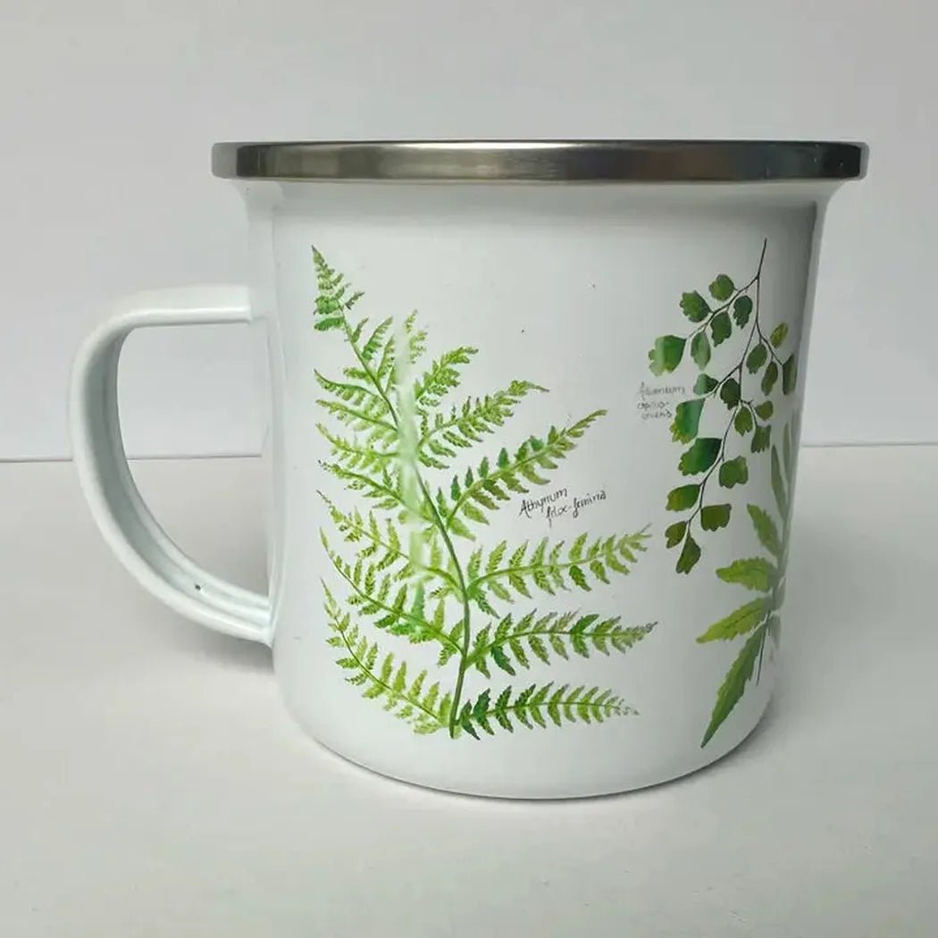 A white Brush & Bark Camping Mug with green fern designs sits on a white surface, showing its handle and metal rim.