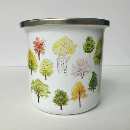 A white Brush & Bark Camping Mug with colorful tree illustrations on it sits on a white surface against a white background.