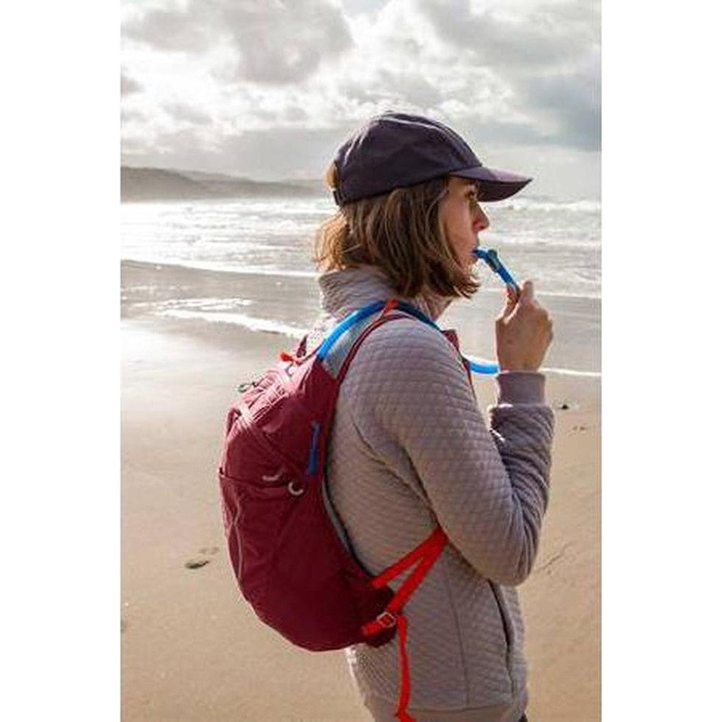 A woman on a beach wears a CamelBak Big Bite Valve with a red backpack and gray jacket.