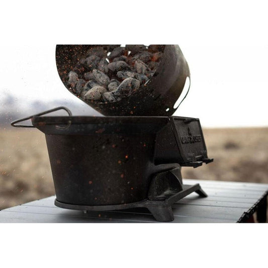 A top-down view of the Camp Chef Cast Iron Charcoal Grill with charcoal being poured into it on a wooden surface.