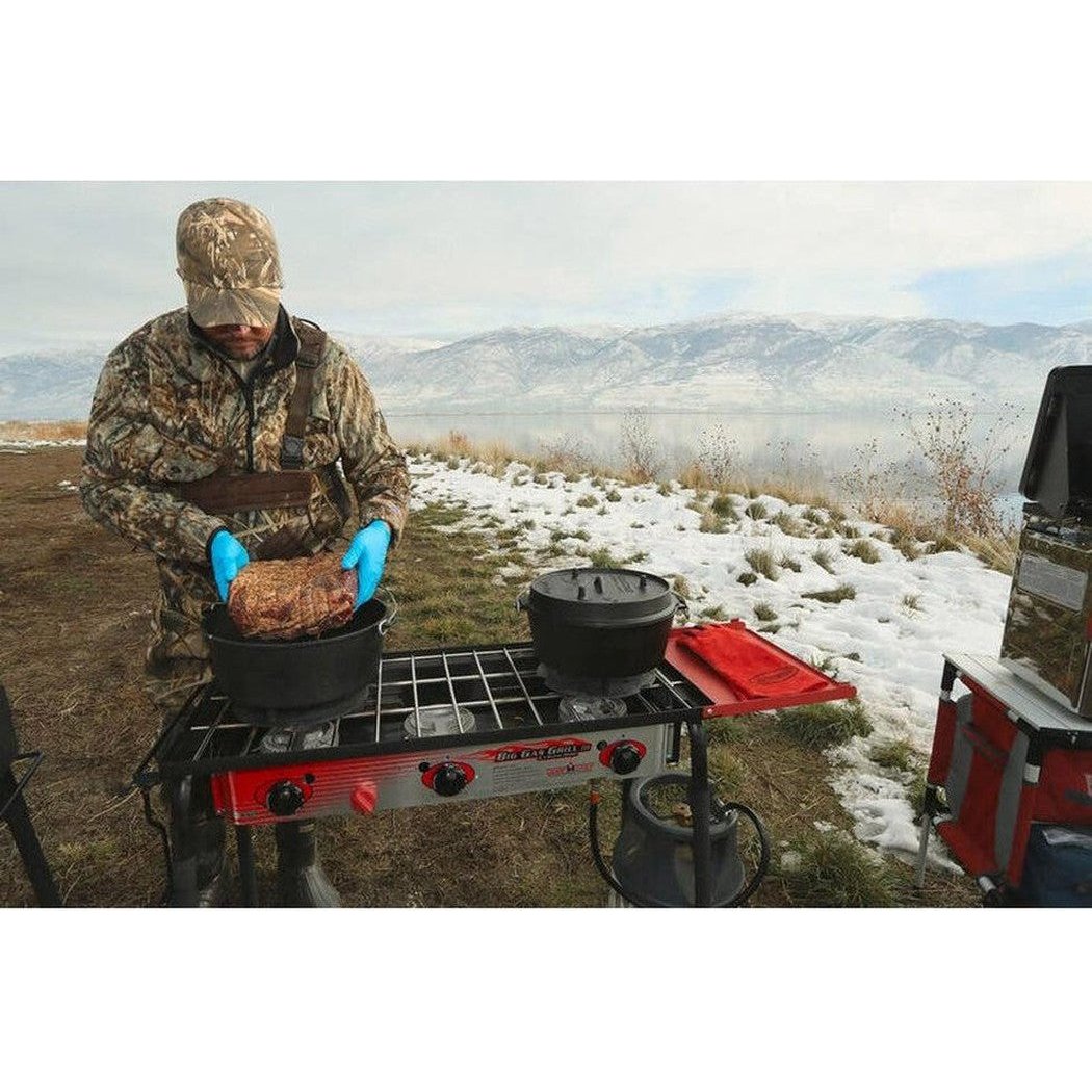 A man in camouflage prepares food with a Camp Chef Cast Iron Deluxe Dutch Oven 14" on a portable stove in a snowy, mountainous outdoor setting.