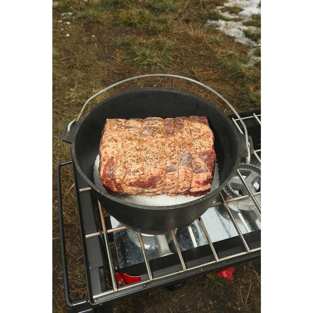 A Camp Chef Cast Iron Deluxe Dutch Oven 14" sits on a portable grill outdoors with a piece of seasoned meat cooking inside.