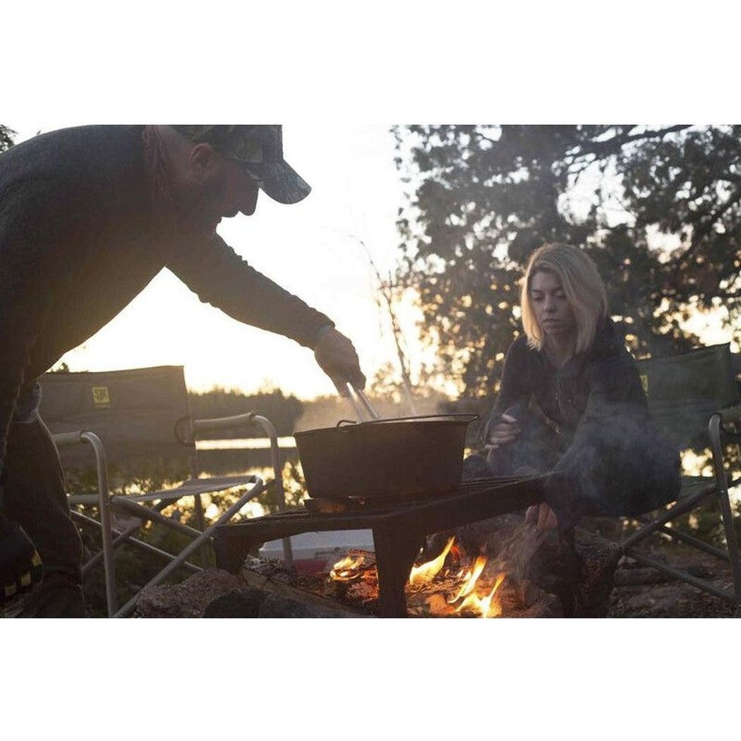 A man stirs a Camp Chef Cast Iron Deluxe Dutch Oven 14" over a campfire while a woman sits nearby in a wooded setting.