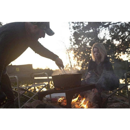 A man stirs a Camp Chef Cast Iron Deluxe Dutch Oven 14" over a campfire while a woman sits nearby in a wooded setting.