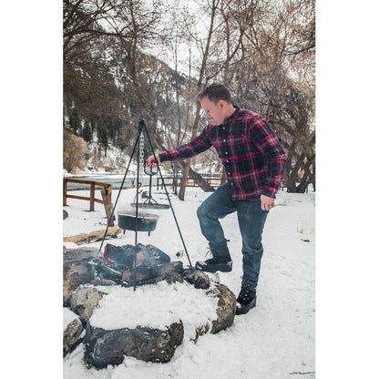 A man in a plaid shirt cooks with a Camp Chef Dutch Oven Tripod over a snowy campfire in a wooded area.