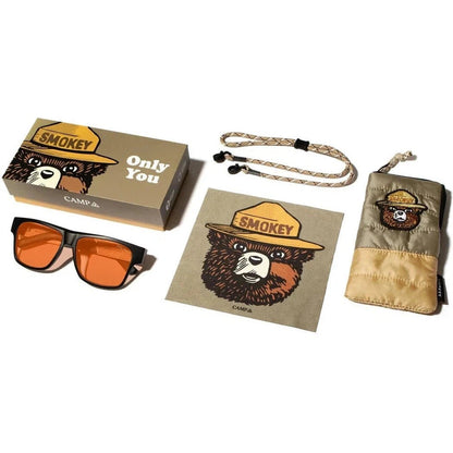 A top-down view of the Camp Eyewear Cliff - Smokey Bear sunglasses in Matte Black with orange lenses, a matching Smokey Bear box, a cleaning cloth, and a case, all on a white background.