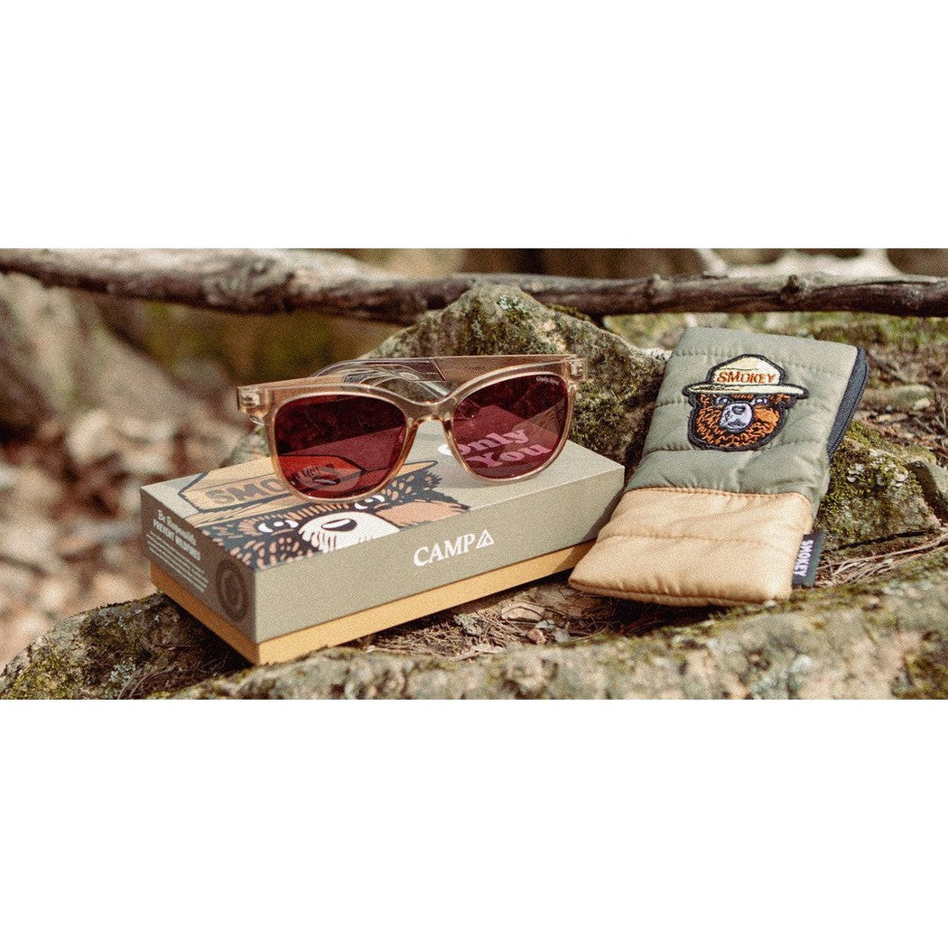 A pair of Camp Eyewear Cove - Smokey Bear sunglasses in Desert color sit on a box with a Smokey Bear case next to them on a rock.
