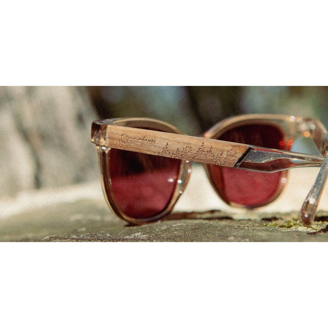 Side view of the Camp Eyewear Cove - Smokey Bear sunglasses with a wooden arm and red lenses on a stone surface.