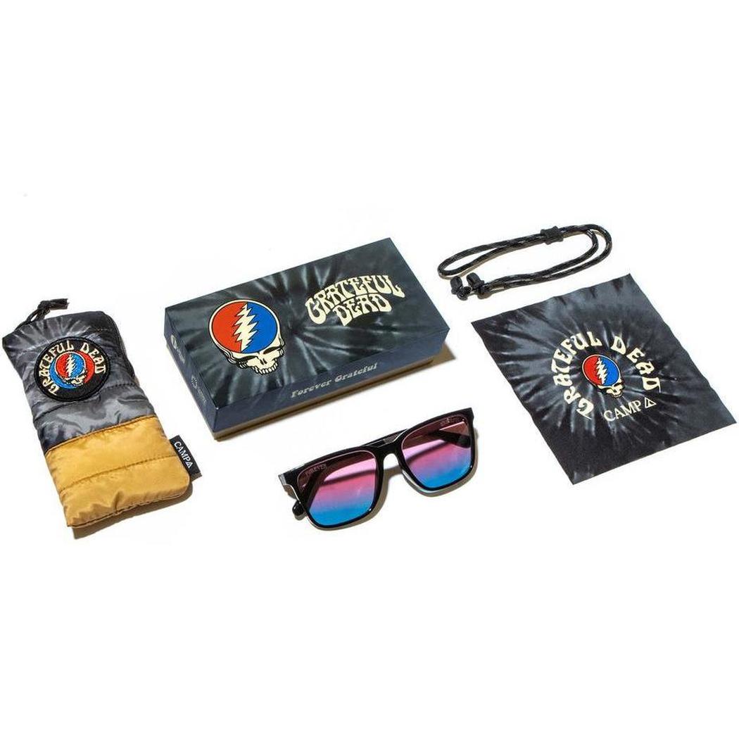 A top-down view of the Camp Eyewear Crag: Grateful Dead Stealie Edition sunglasses with black frames and blue-purple lenses, alongside a branded case, cloth, and lanyard on a white background.