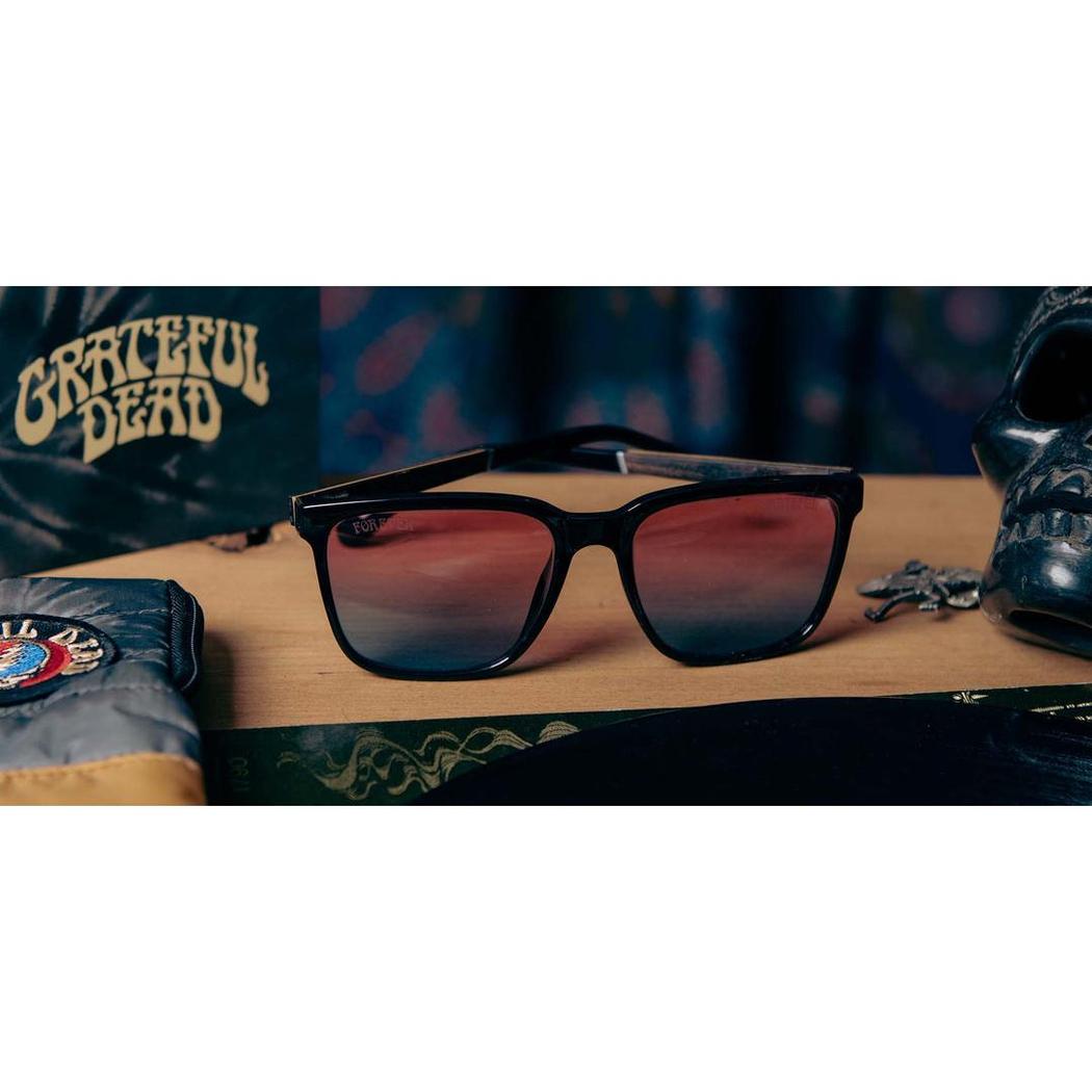 A pair of black Camp Eyewear Crag sunglasses with red-tinted lenses sit on a wooden surface with a Grateful Dead shirt and a skull in the background.