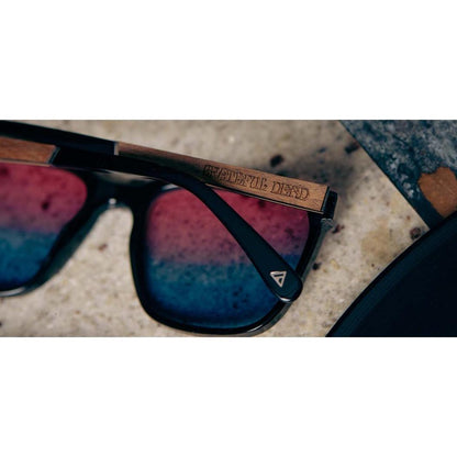 A close-up of the Camp Eyewear Crag: Grateful Dead Stealie Edition sunglasses with black frames and a wooden temple engraved with "Grateful Dead" on a concrete surface.