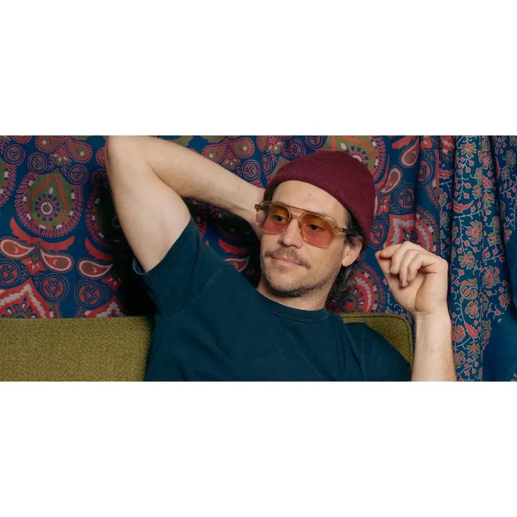 A man wearing the Camp Eyewear Glacier: Grateful Dead Bears Edition sunglasses relaxes on a couch with a colorful patterned background.