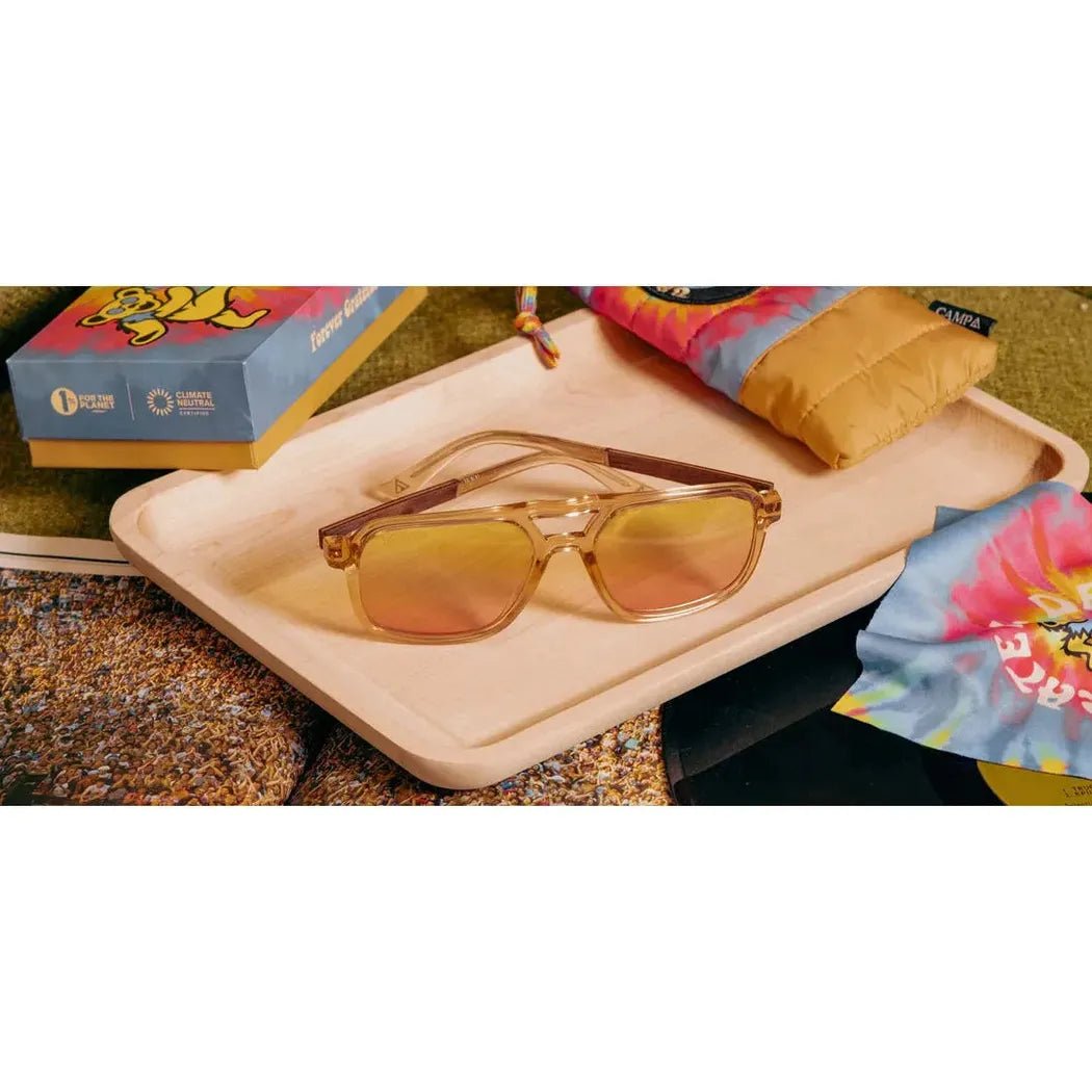 A pair of Camp Eyewear Glacier: Grateful Dead Bears Edition sunglasses with desert colored frames and engraved details rest on a light wooden tray with a colorful tie-dye cloth nearby.