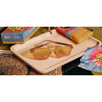 A pair of Camp Eyewear Glacier: Grateful Dead Bears Edition sunglasses with desert colored frames and engraved details rest on a light wooden tray with a colorful tie-dye cloth nearby.