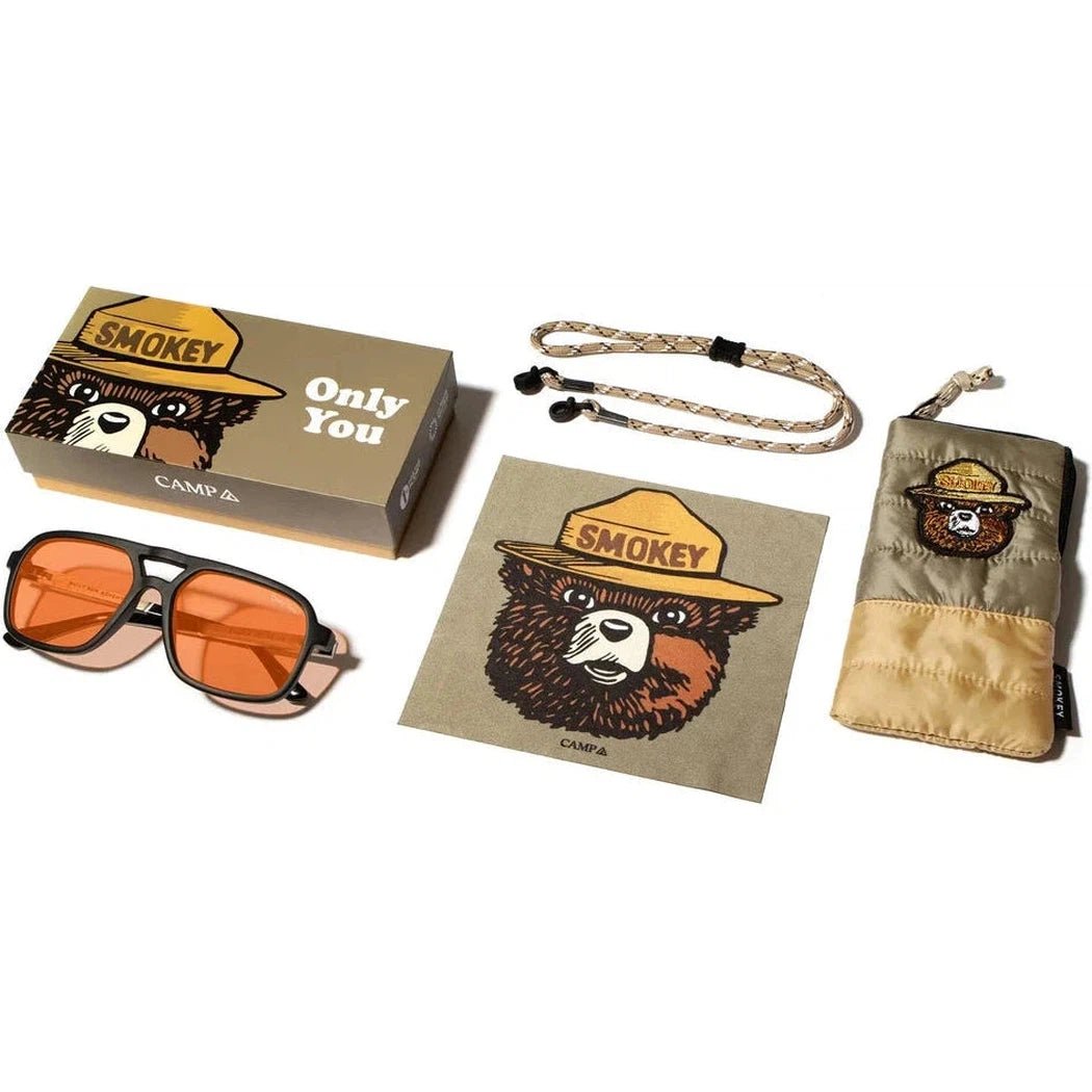 A top-down view of the Camp Eyewear Glacier - Smokey Bear sunglasses in Matte Black with orange lenses, a Smokey Bear box, a matching pouch, and a lanyard.