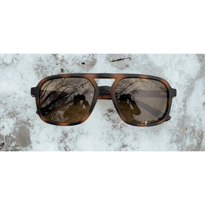 The Camp Eyewear Glacier sunglasses with Black // Walnut frames rest on a snowy surface, showing reflections of trees and a photographer.
