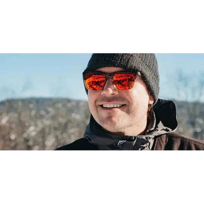 A man wears the Camp Eyewear Ridge sunglasses in Black // Ebony, smiling at a snowy outdoor scene.