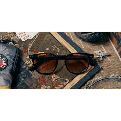 Black Camp Eyewear Topo: Grateful Dead Stealie Edition sunglasses rest on a patterned surface with a wallet and keys nearby.