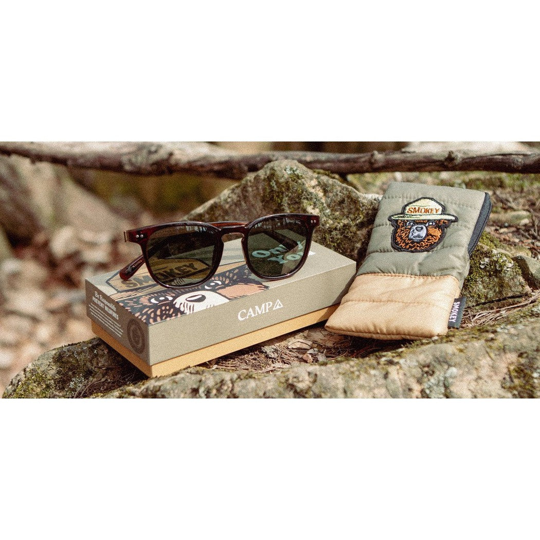 A pair of Camp Eyewear Topo - Smokey Bear sunglasses in Clay rest on their box and case atop a rock in a wooded setting.