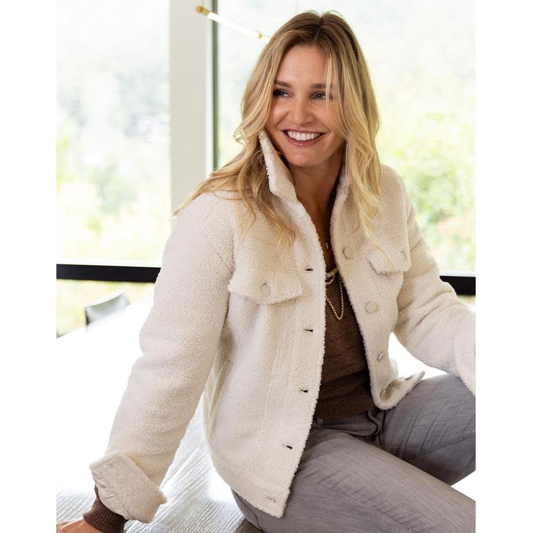 A woman wearing the Carve Designs Women's Rhea Boucle Trucker jacket in Birch sits by a window with greenery outside.
