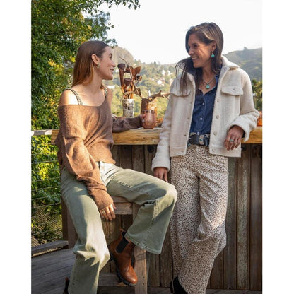Two women chat on a wooden deck wearing Carve Designs Women's Rhea Boucle Trucker jackets in Birch, with a scenic mountain backdrop.