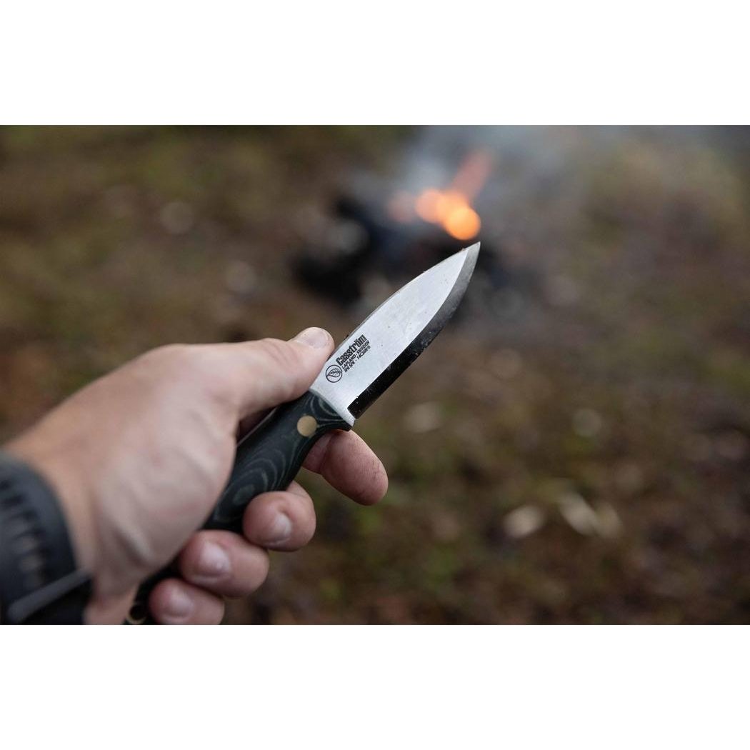 A hand holds the Casstrom No 8 Swedish Forest Knife Green Micarta with Firesteel, showing the blade and handle, with a campfire in the background.
