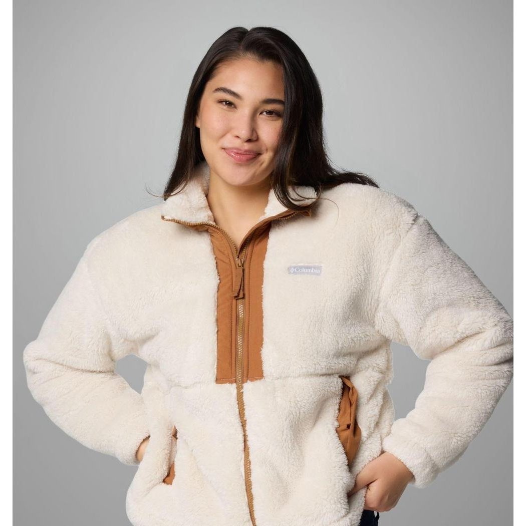 A woman wearing the Columbia Sportswear Women's Boundless Discovery Sherpa Full Zip II Jacket in Chalk/Camel Brown poses with her hands on her hips against a gray background.