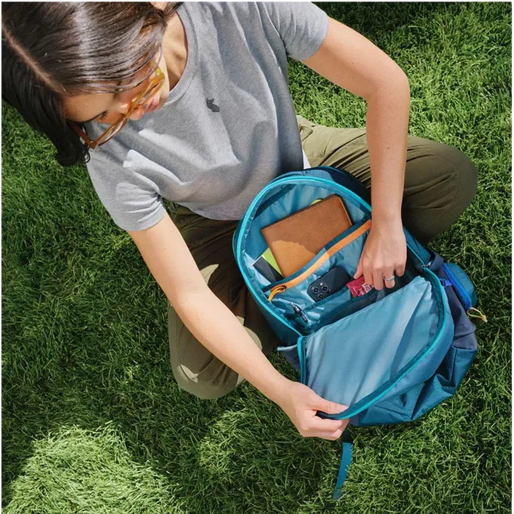 A woman crouches on grass opening the Cotopaxi Allpa 26L Daypack in Blue Spruce/Abyss to show its interior.