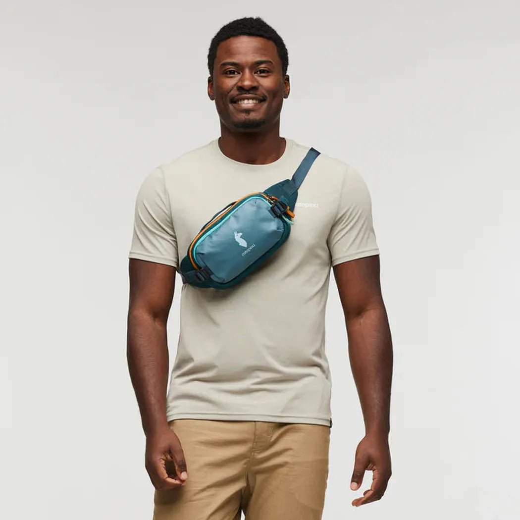 A man wearing the Cotopaxi Allpa X 1.5L Hip Pack in Blue Spruce/Abyss stands against a plain white background.