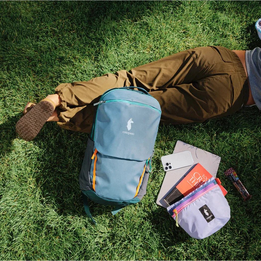 A person lies on green grass with a blue Cotopaxi Cubo Pouch Organizer and a matching backpack nearby.