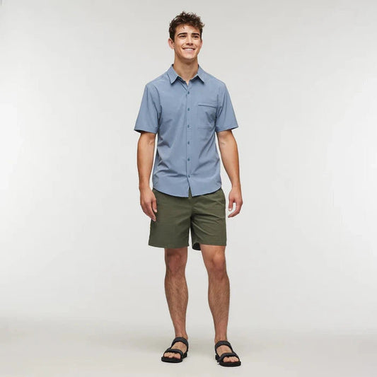 A front view of the Cotopaxi Men's Cambio Button Up in Tempest shows a model wearing it with olive shorts and black sandals against a plain white background.