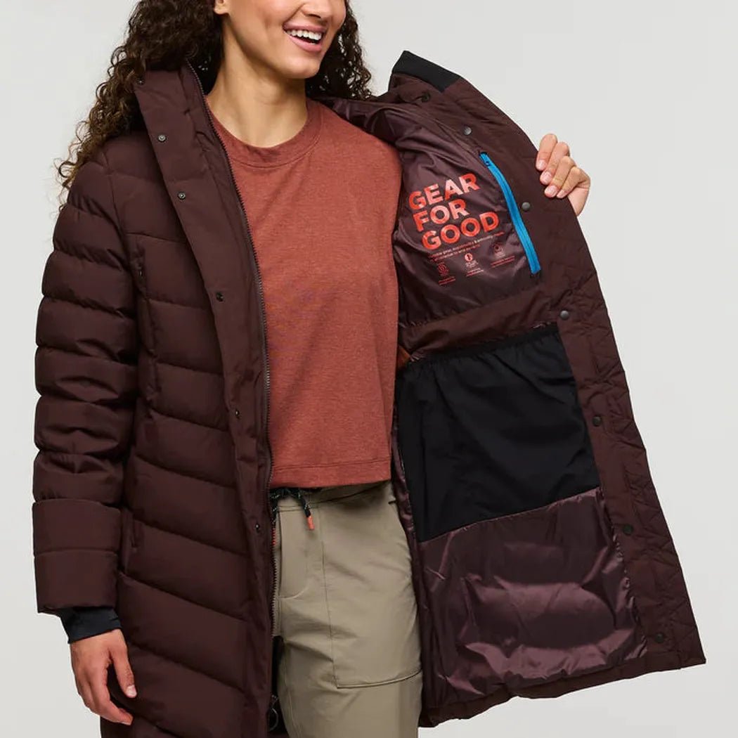 A woman wearing the Cotopaxi Women's Alivio Down Parka in Coffee shows the interior pocket with "Gear for Good" text against a plain background.