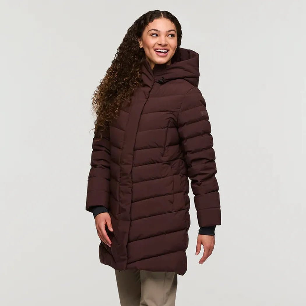 A woman wearing the Cotopaxi Women's Alivio Down Parka in Coffee stands against a plain white background.