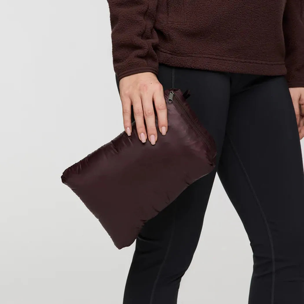 A model wearing the Cotopaxi Women's Fuego Down Parka in Coffee Stripes holds a matching clutch against a plain white background.